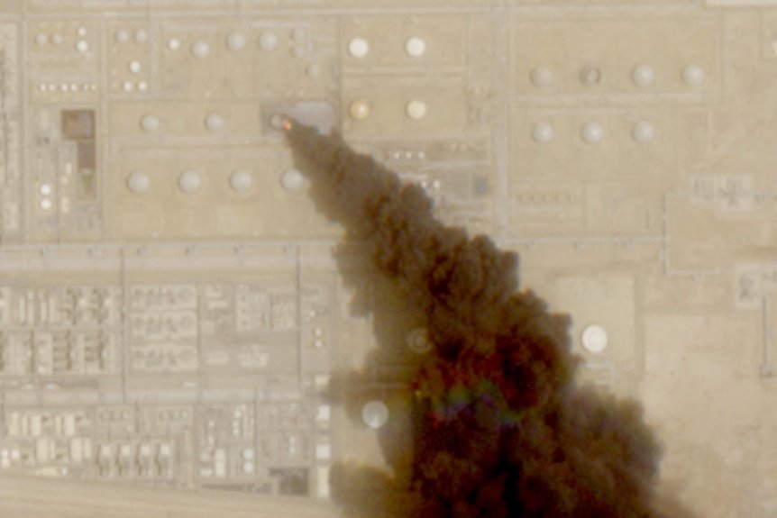 A satellite image of a vast oil facility showing a large plume of smoke burning from a tank.